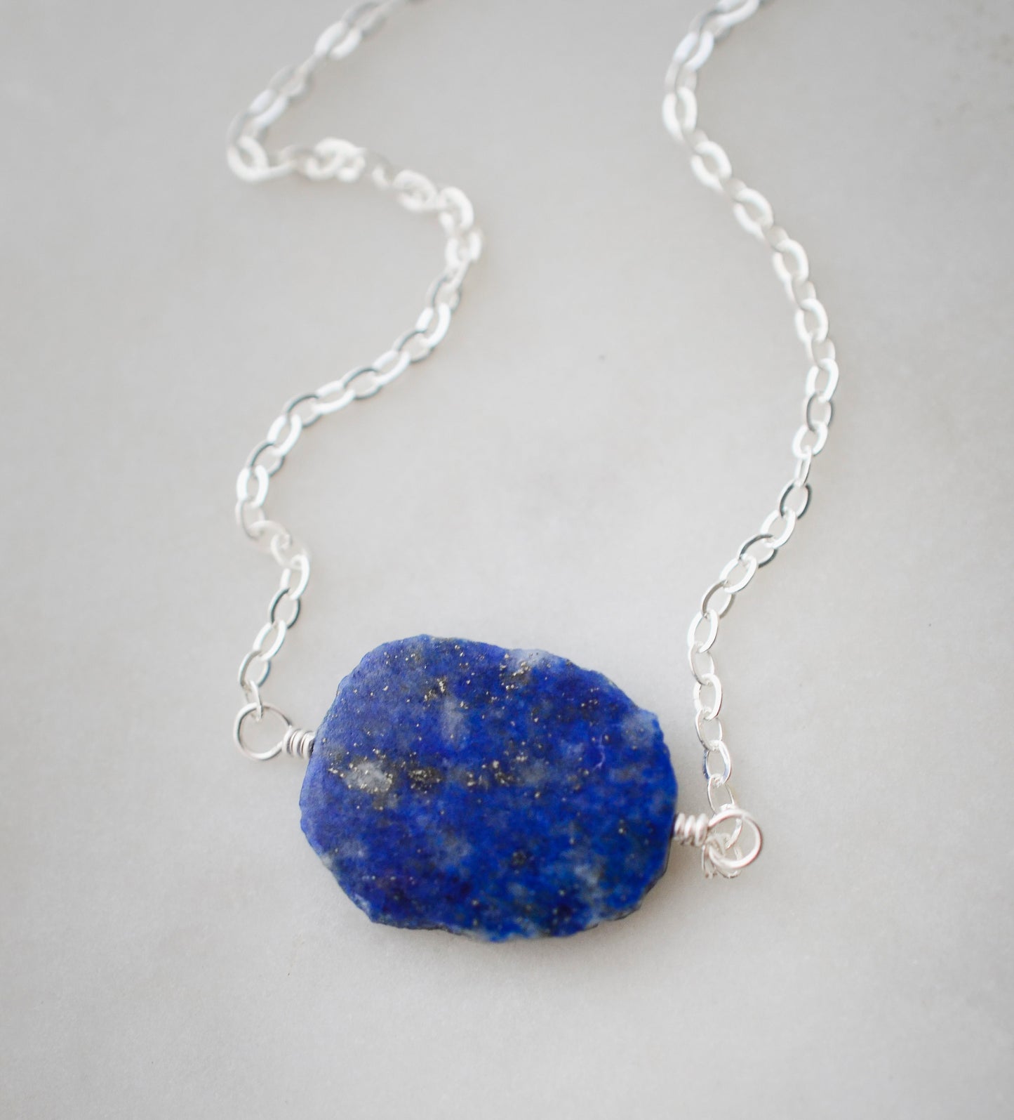 Natural blue lapis lazuli slice set onto a sterling silver chain. The stone is smooth polished with raw edges and oval in shape. Natural pyrite and calcite flecks are visible within each stone.