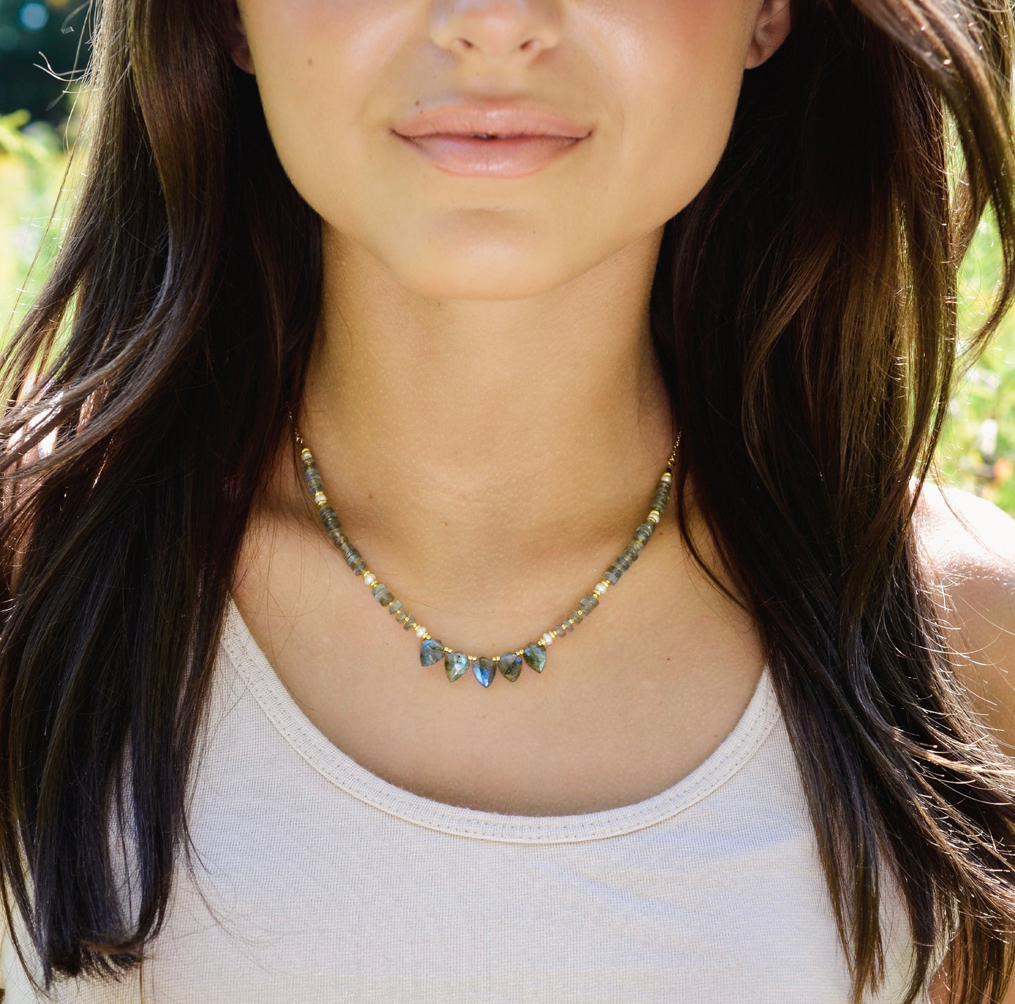 A woman wearing a beaded labradorite and white freshwater pearl necklace with five teardrops at the center. Shown in gold.