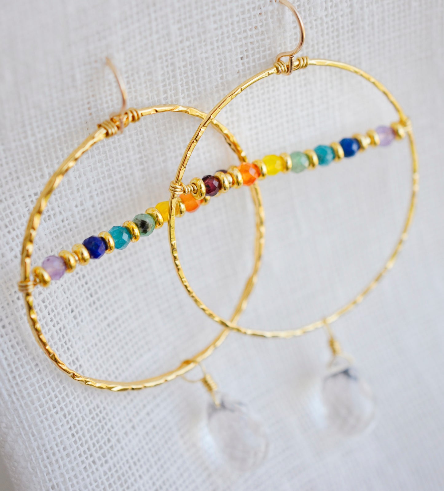 Top View. Large hammered hoop earrings with a bar of rainbow crystals spanning its center. The hoop is textured and has natural clear crystal quartz teardrop dangles. The gold style is shown.