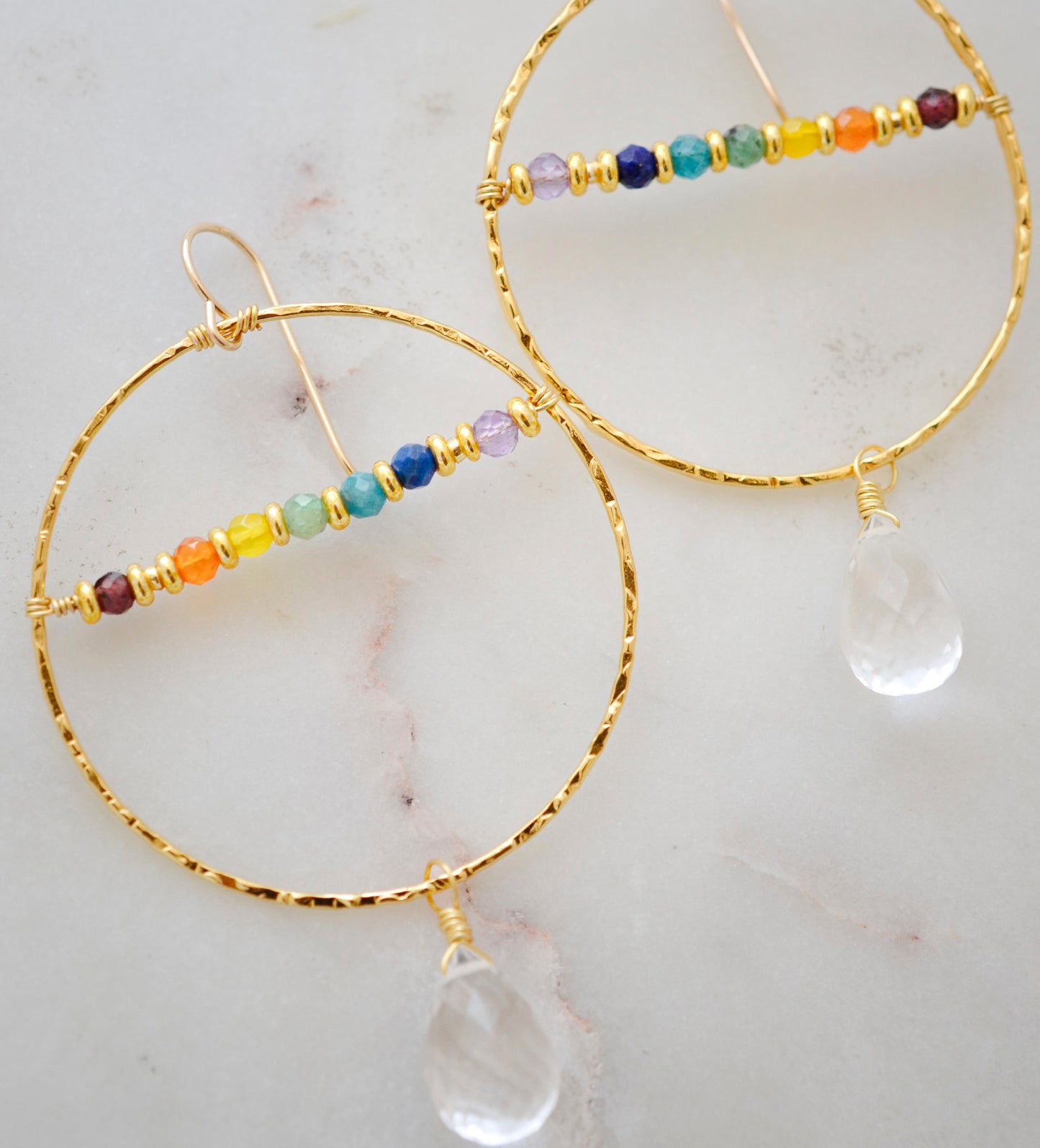 Large hammered hoop earrings with a bar of rainbow crystals spanning its center. The hoop is textured and has natural clear crystal quartz teardrop dangles. The gold style is shown.