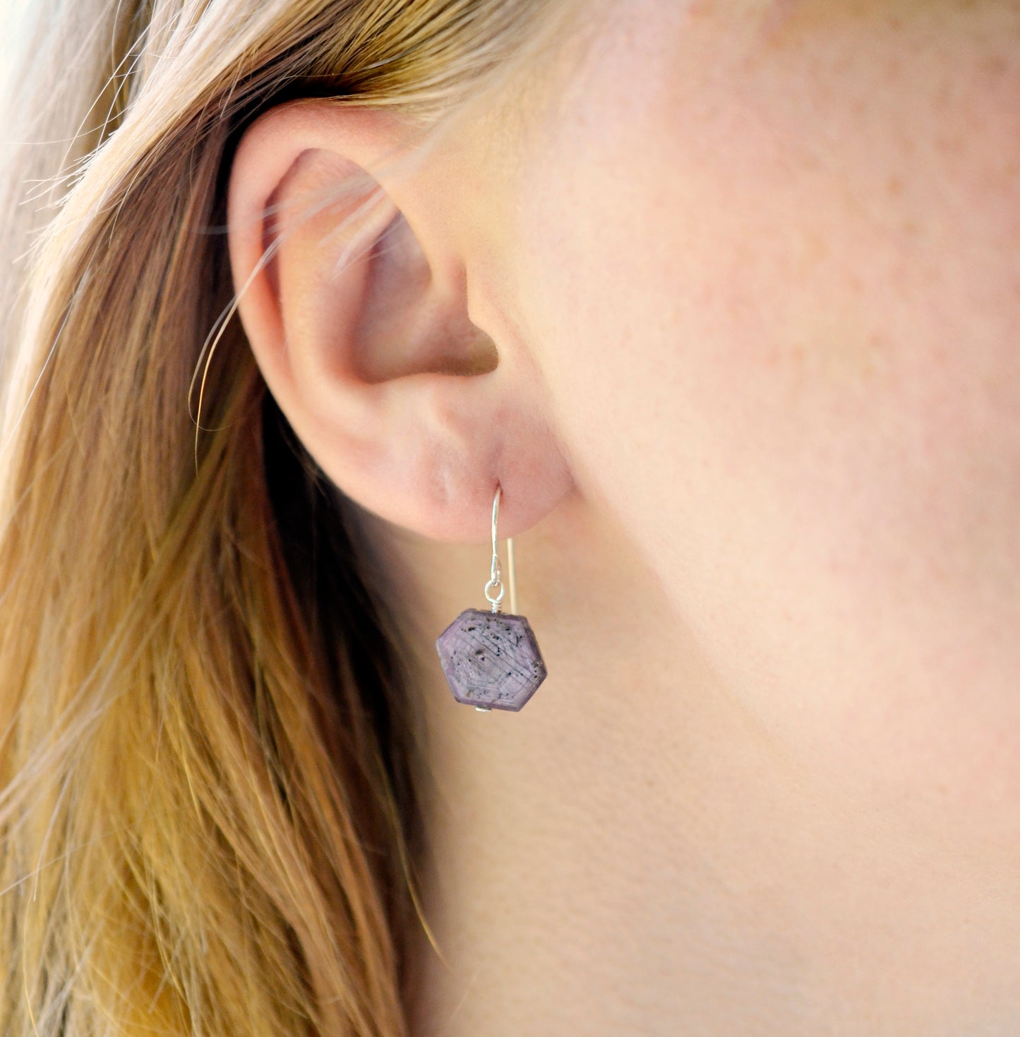 Purple-pink raw ruby stones suspended from sterling silver earwires. The stones are rough and in their natural hexagonal shape. Modeled image.