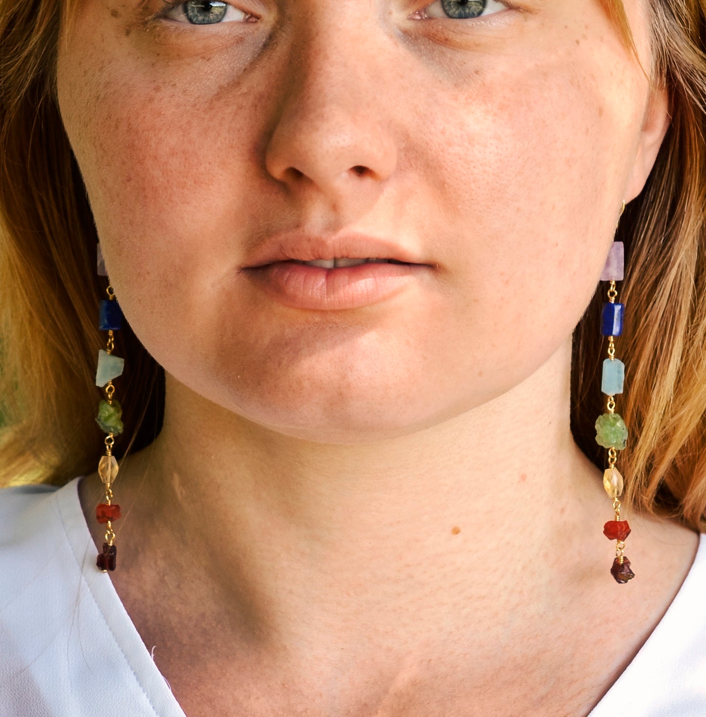 Long gemstone earrings made with a rainbow of raw gemstones. Colors include: purple, blue, aqua, green, yellow, orange, and red. Modeled image.