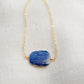 Natural blue lapis lazuli slice set onto a 14k gold filled chain. The stone is smooth polished with raw edges and oval in shape. Natural pyrite and calcite flecks are visible within each stone. 