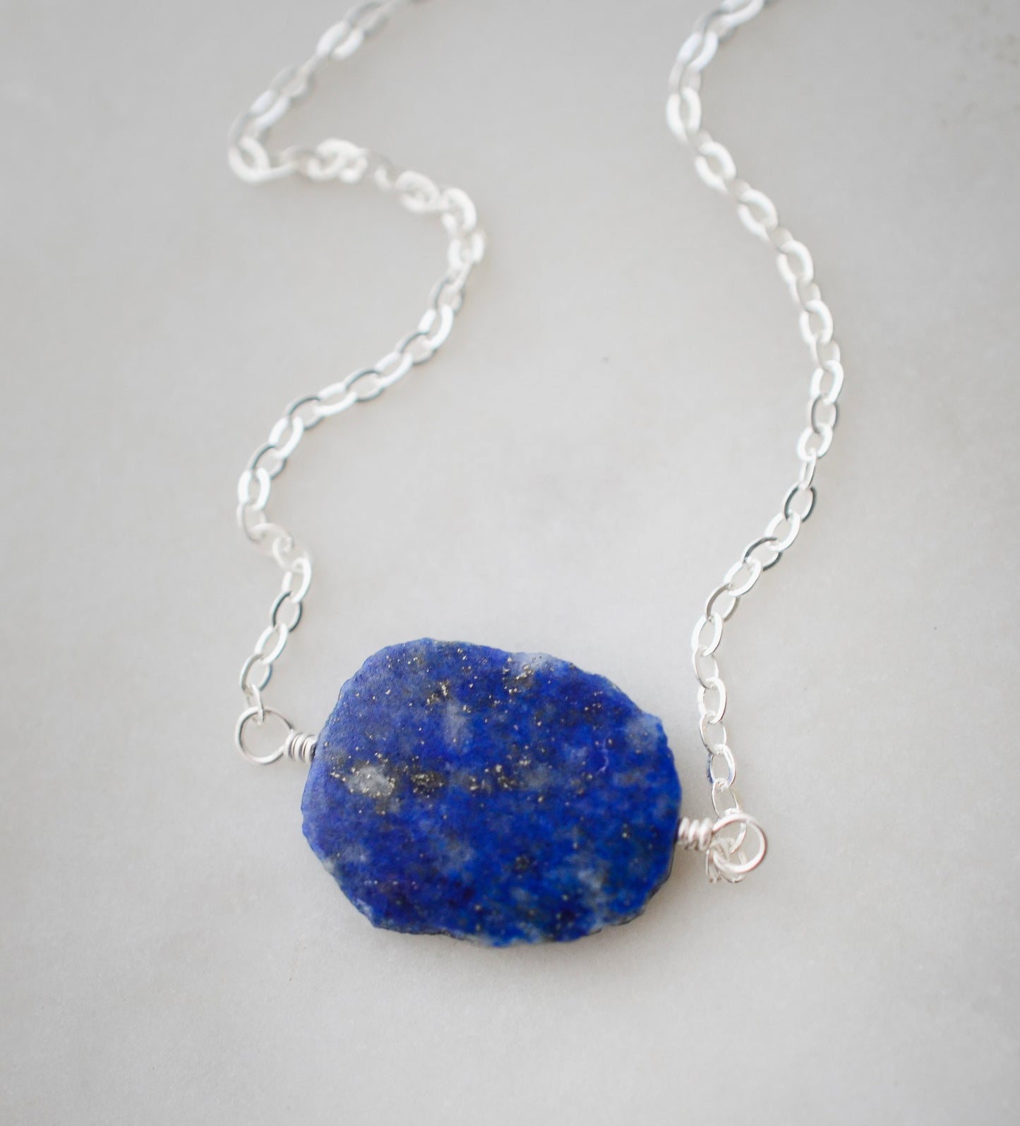 Natural blue lapis lazuli slice set onto a sterling silver chain. The stone is smooth polished with raw edges and oval in shape. Natural pyrite and calcite flecks are visible within each stone. 