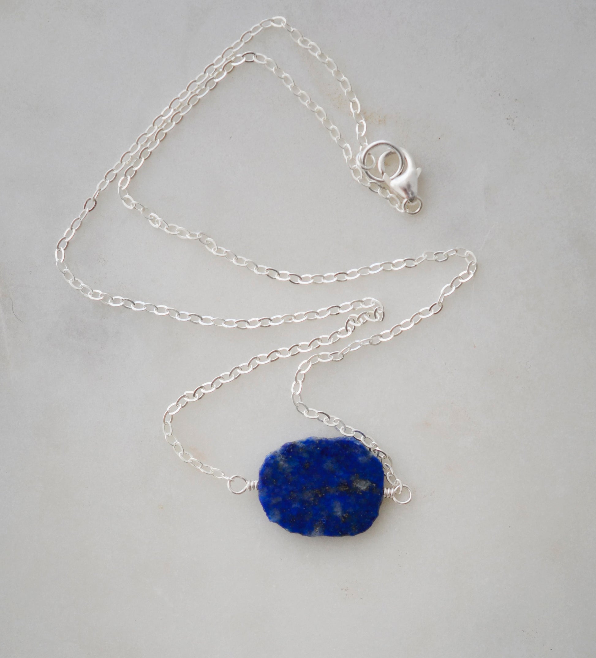 Natural blue lapis lazuli slice set onto a sterling silver chain. The stone is smooth polished with raw edges and oval in shape. Natural pyrite and calcite flecks are visible within each stone. 