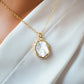 A smooth polished natural, white mother of pearl slice has been set in a 24k gold electroplated bezel and suspended from a 14k gold filled chain. The shell shimmers in shades of white, silver, and pink. It is shown on white fabric.