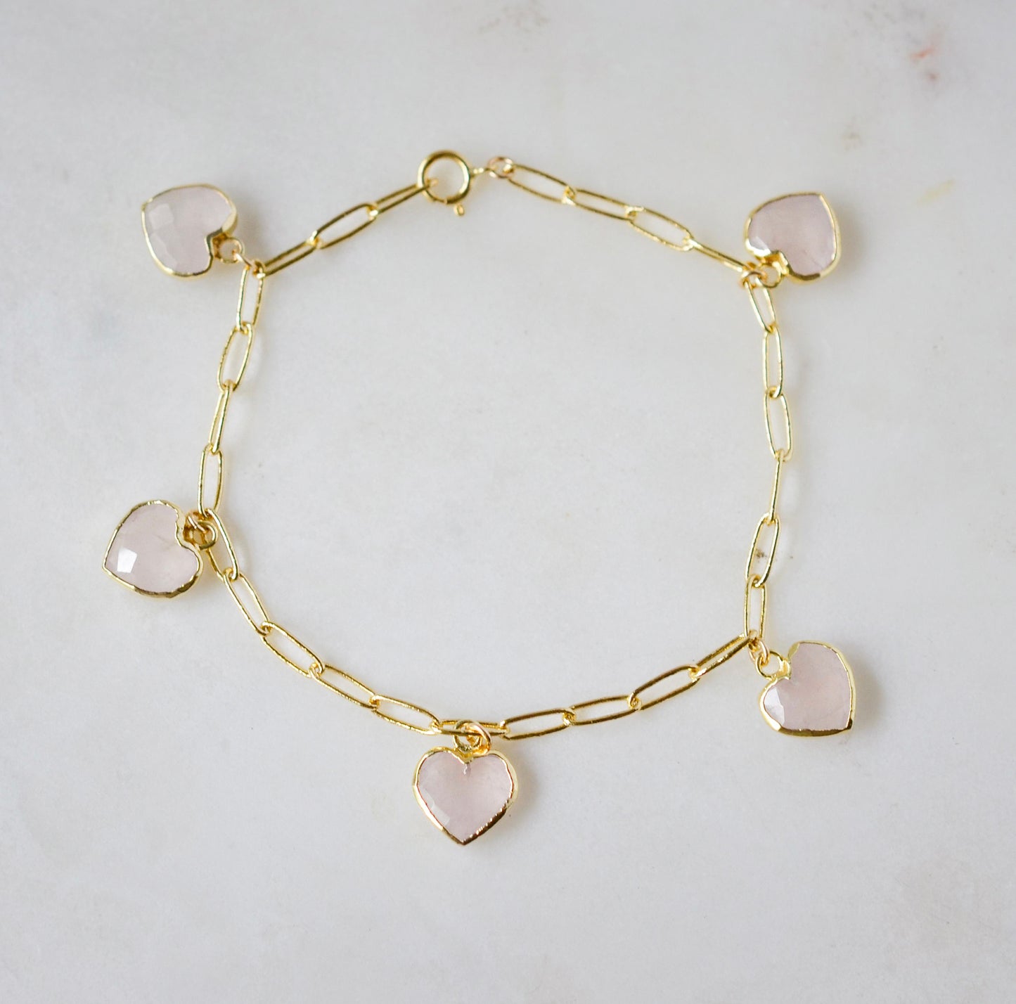 A gold paperclip chain bracelet with heart-shaped pink rose quartz gemstones on a light gray background. 