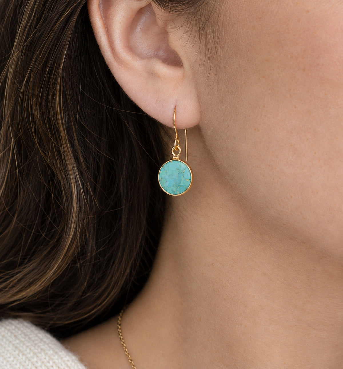 A close-up of a woman's ear wearing a gold dangle earring with a round turquoise-blue stone. Handmade with natural smooth polished turquoise by Gemnia in Connecticut. 