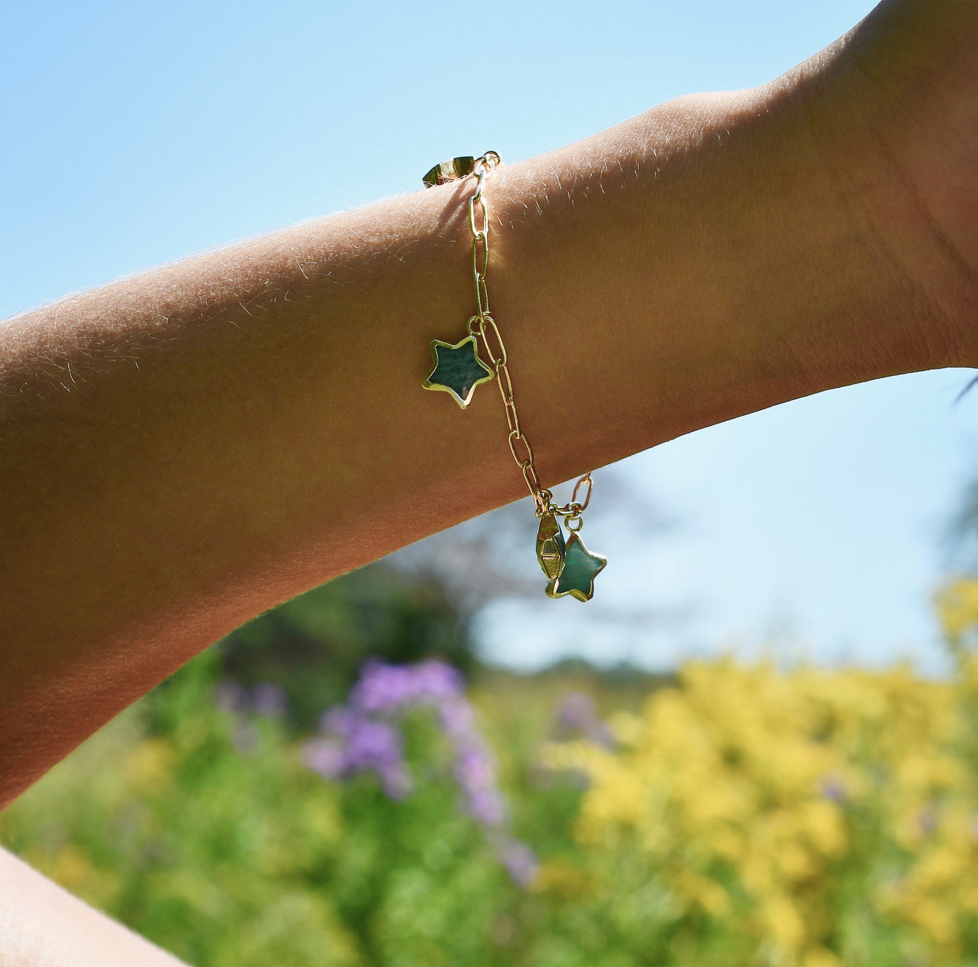 Gold bracelet with Amazonite gemstone star-shaped charms on a wrist against a blurred natural background.