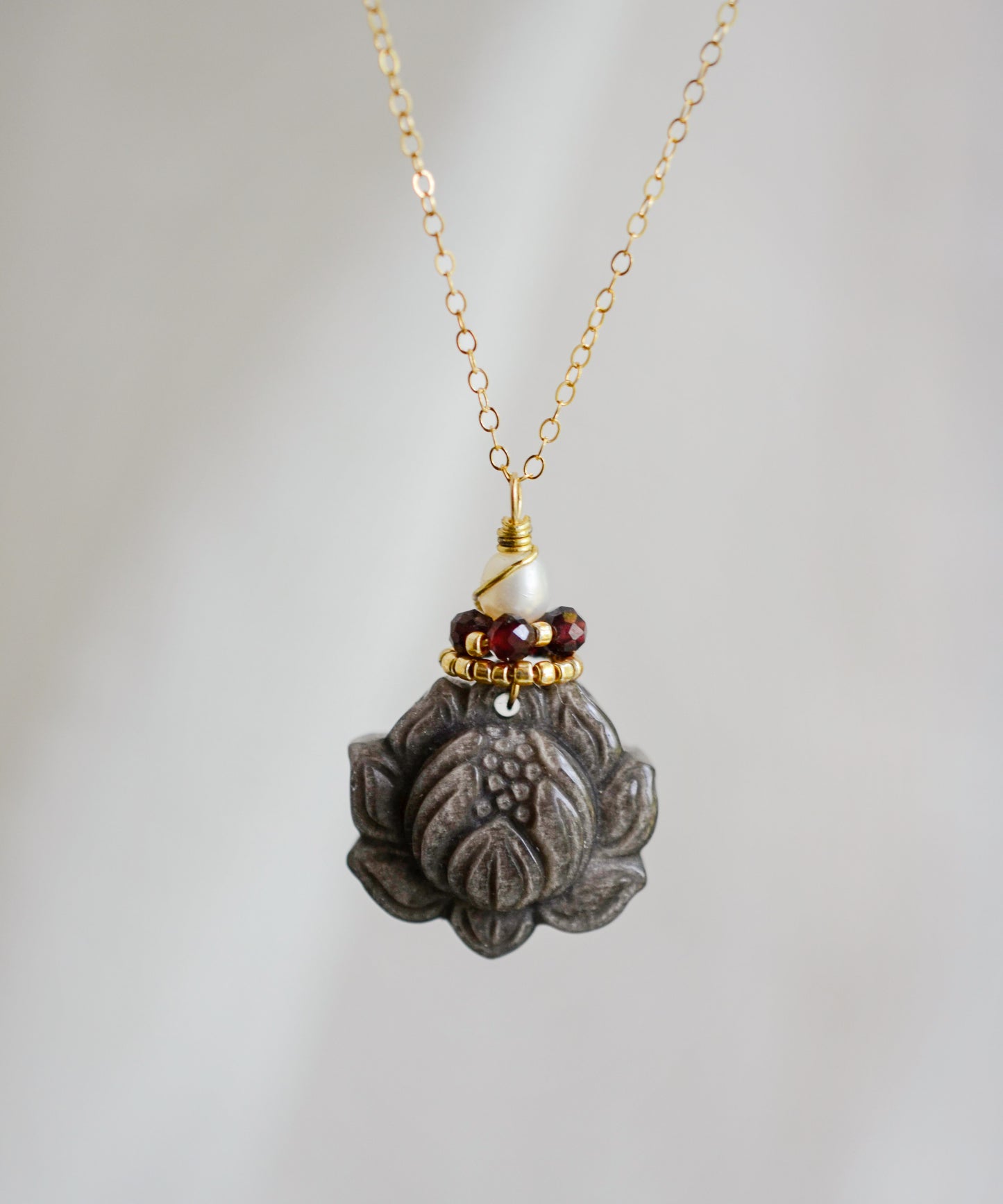 A handmade gemstone pendant featuring a carved obsidian lotus. Garnet, pearl, and gold beads decorate its top. The chain is 14k gold filled.