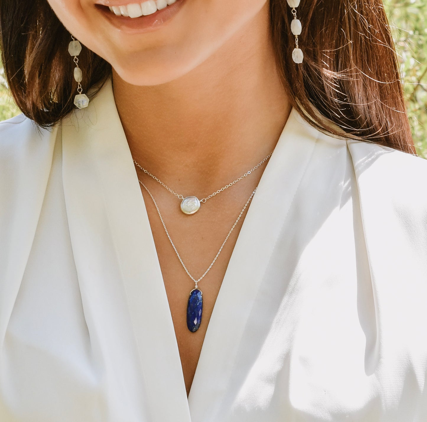 A modeled image of a woman wearing two layered necklaces. One is a white freshwater pearl. The other is a real blue lapis lazuli faceted oval stone pendant. Shown in sterling silver. This jewelry is handmade by Gemnia.