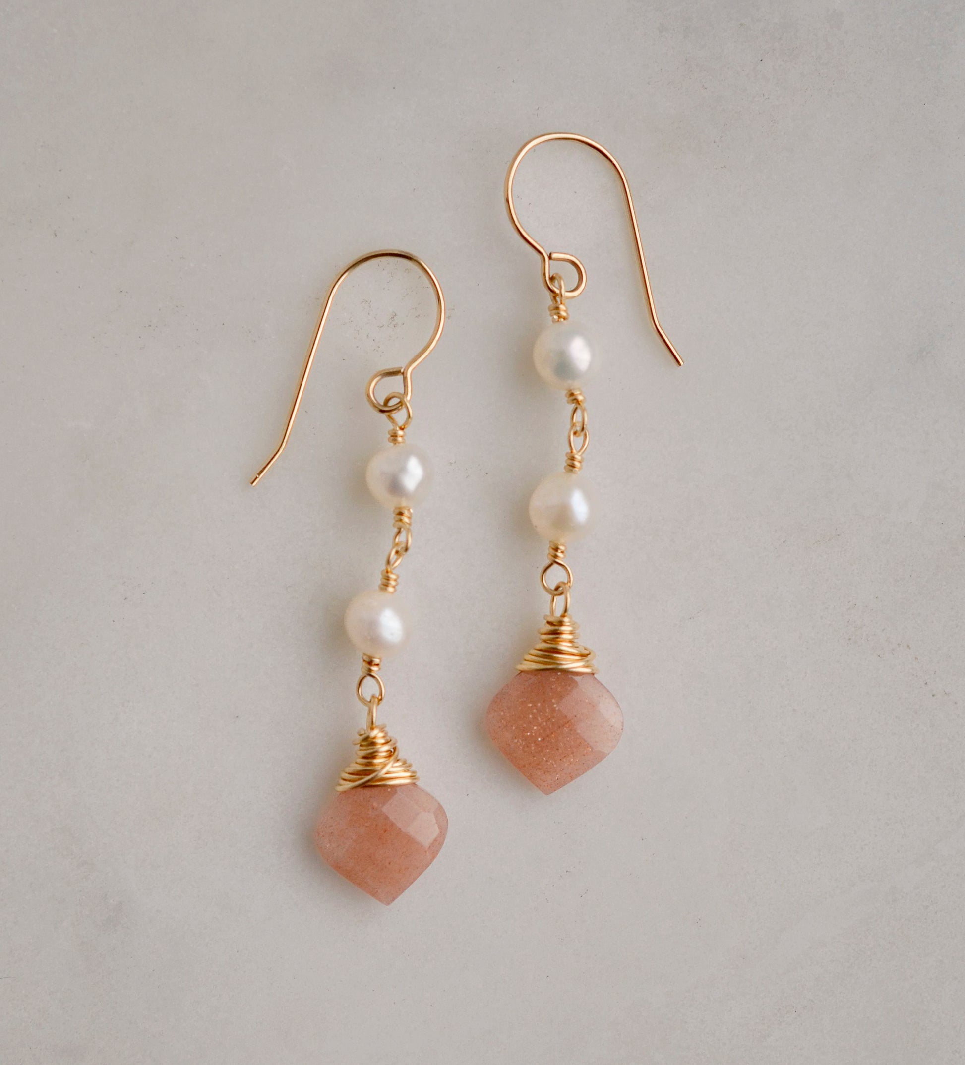 Two white semi-round pearls hang over natural peach Moonstone faceted drops. The gold style is shown.