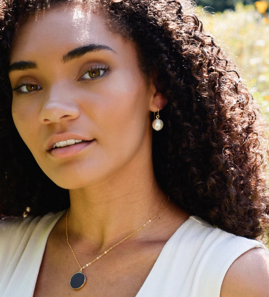 Natural white freshwater pearl dangle earring. The pearl is set in 22k gold electroplate and suspended from 14k gold filled earwires. Modeled image. Also shown, black onyx necklace.