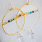 Top View. Large hammered hoop earrings with a bar of rainbow crystals spanning its center. The hoop is textured and has natural clear crystal quartz teardrop dangles. The gold style is shown.