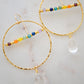 Large hammered hoop earrings with a bar of rainbow crystals spanning its center. The hoop is textured and has natural clear crystal quartz teardrop dangles. The gold style is shown.