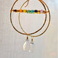 Large hammered hoop earrings with a bar of rainbow crystals spanning its center. The hoop is textured and has natural clear crystal quartz teardrop dangles. The gold style is shown.