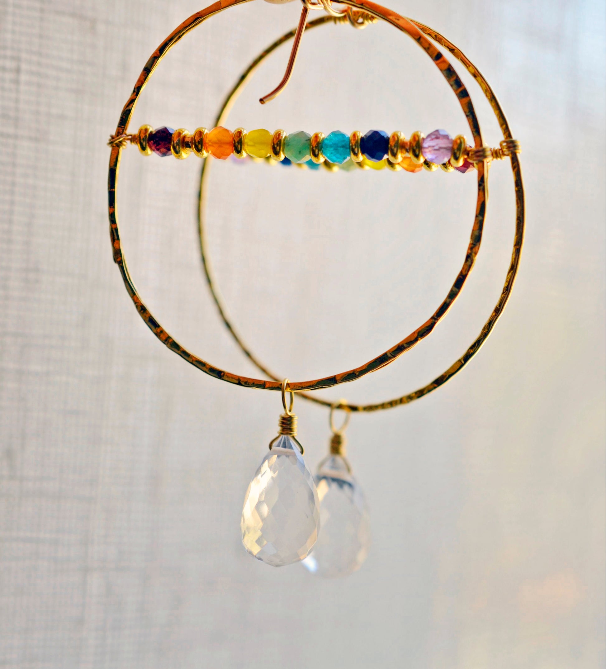 Large hammered hoop earrings with a bar of rainbow crystals spanning its center. The hoop is textured and has natural clear crystal quartz teardrop dangles. The gold style is shown.