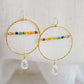 Large hammered hoop earrings with a bar of rainbow crystals spanning its center. The hoop is textured and has natural clear crystal quartz teardrop dangles. The gold style is shown.