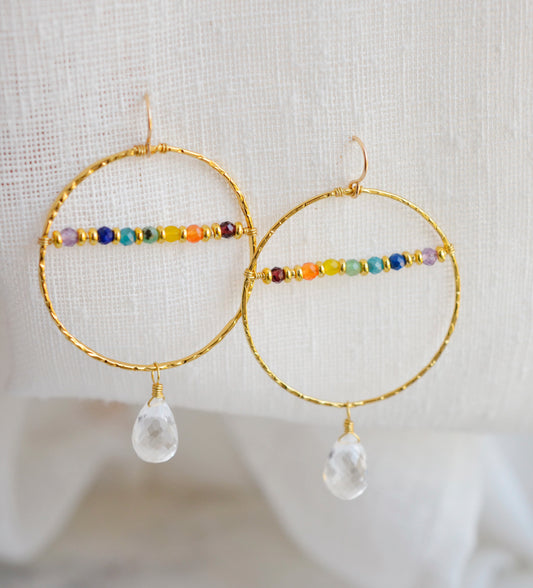 Large hammered hoop earrings with a bar of rainbow crystals spanning its center. The hoop is textured and has natural clear crystal quartz teardrop dangles. The gold style is shown.