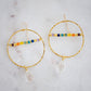 Large hammered hoop earrings with a bar of rainbow crystals spanning its center. The hoop is textured and has natural clear crystal quartz teardrop dangles. The gold style is shown.