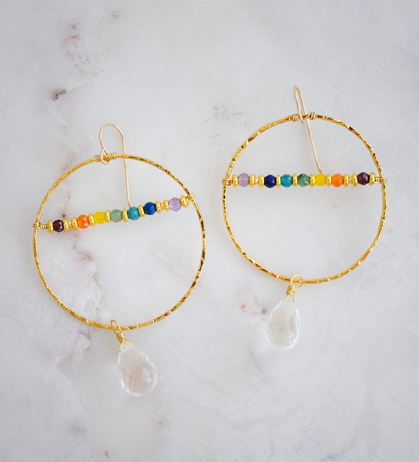 Large hammered hoop earrings with a bar of rainbow crystals spanning its center. The hoop is textured and has natural clear crystal quartz teardrop dangles. The gold style is shown.