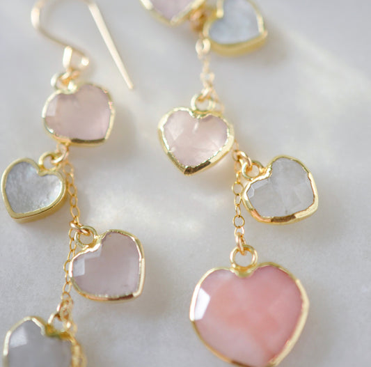 Long pink stone heart earrings in 14k gold filled. There are five heart crystal dangles hanging from a simple chain forming a cluster. The stones include: rose quartz, mother of pearl, pink opal, and rainbow moonstone. They are approximately three inches long.
