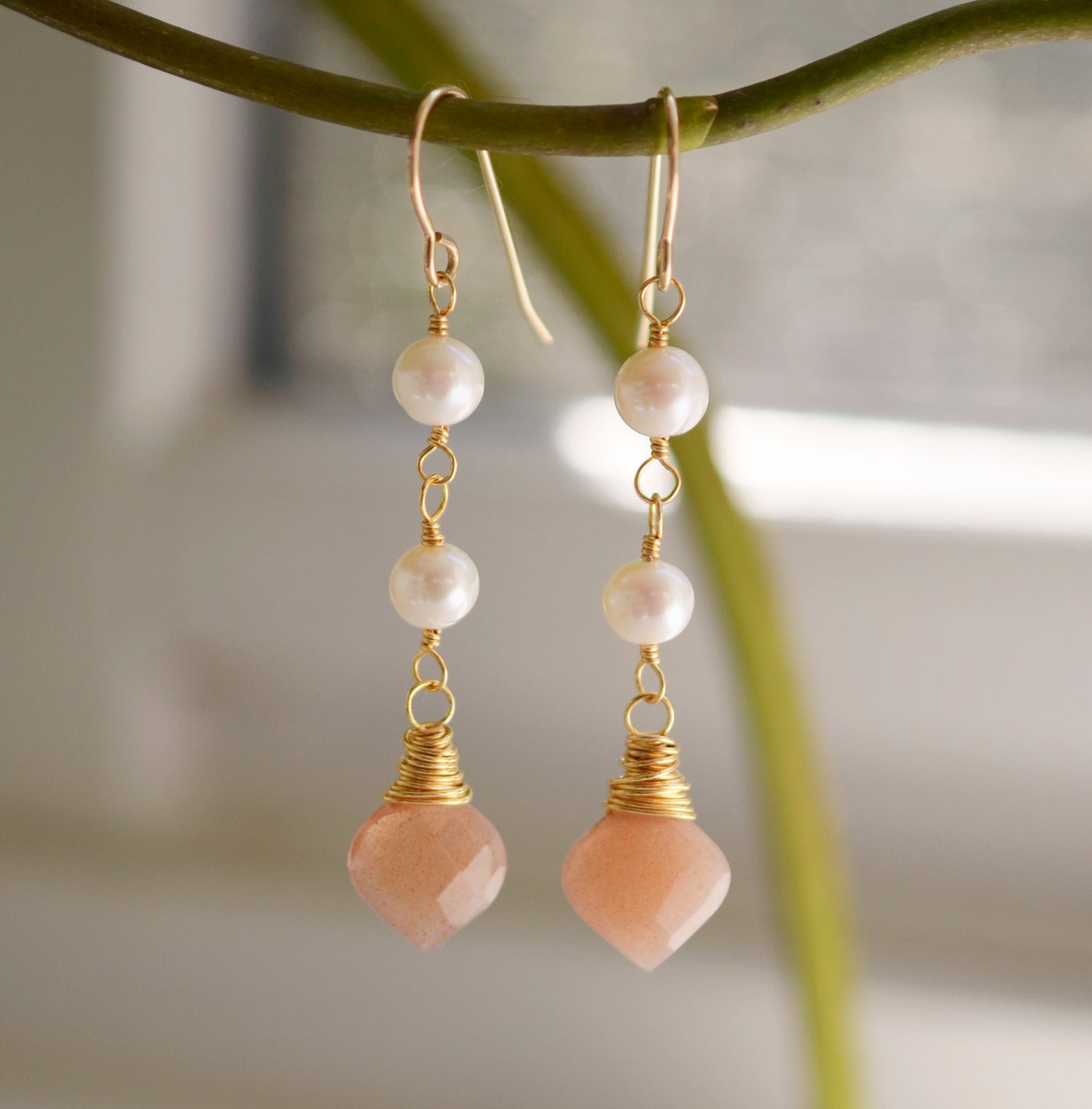 Two white semi-round pearls hang over natural peach Moonstone faceted drops. The gold style is shown.