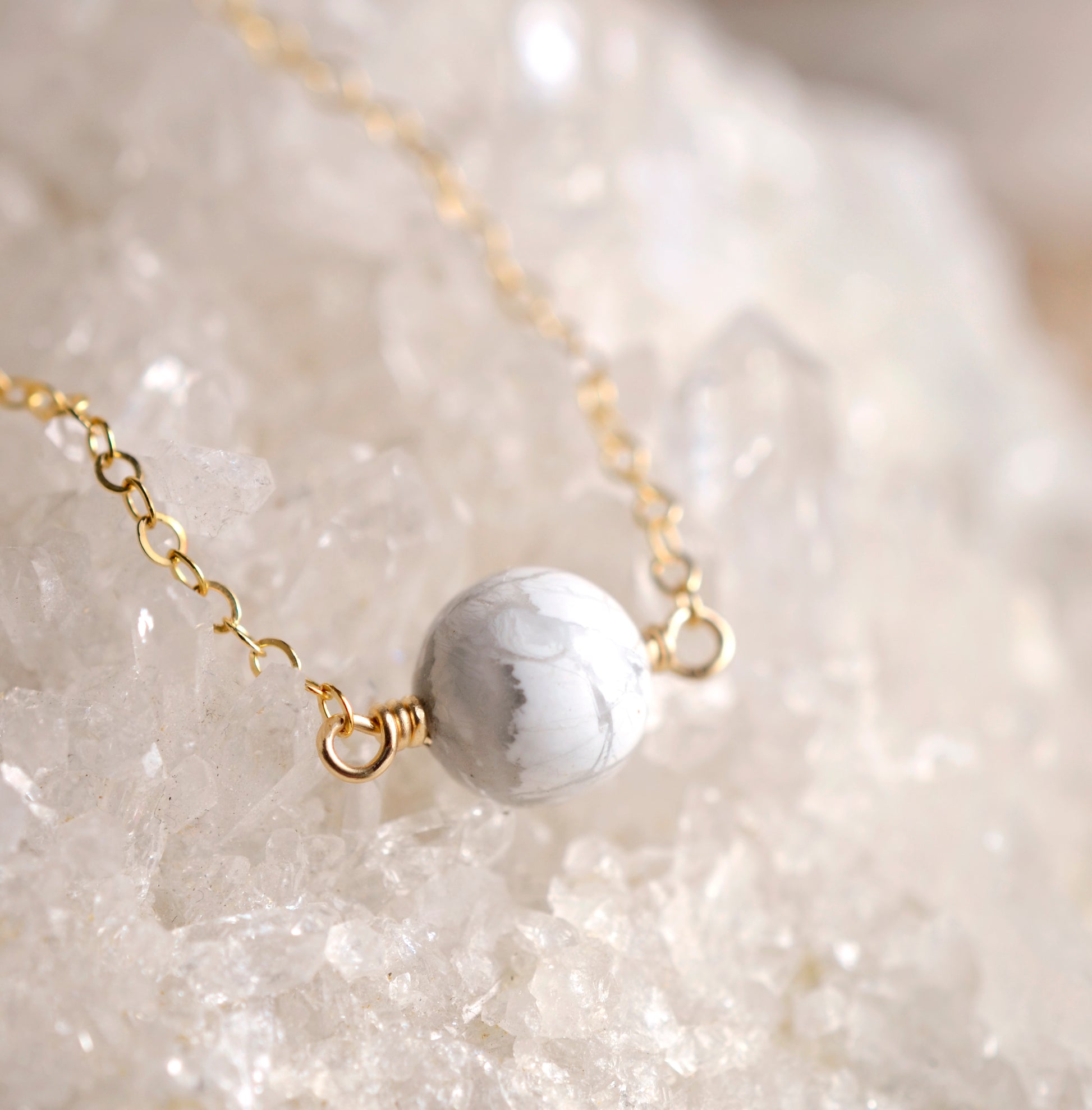 Close up image. A natural white howlite necklace on a crystal quartz background. The stone is round with natural gray marbling and set on a 14k gold filled chain. Also available in sterling silver. 